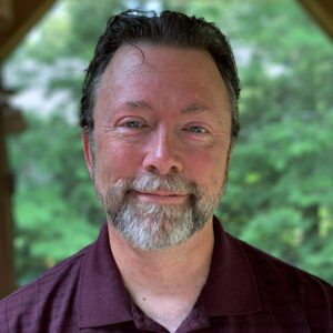 Headshot of 60 year white male with salt and pepper beard and dark hair, smiling at camera